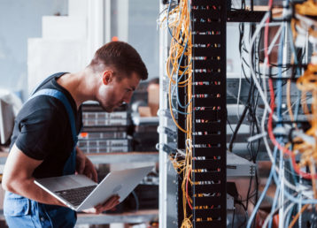 Young man in uniform and with laptop works with internet equipment and wires in server room